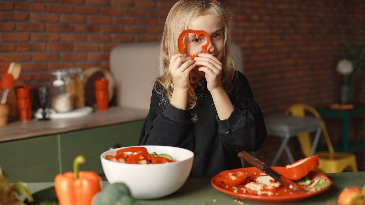 Funny cheerful girl looking through slice of pepper while preparing healthy vegetarian salad at table with plate with knife and salad bowl